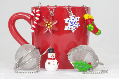 six tea ball infusers shown on a red mug, each with a different Christmas ornament attached to its chain: Snowman with black hat, green holly leaf with red berry, red and white candy cane, red poinsettia, white snowflake with blue decorative dots, red stocking with green heal and toe with yellow top.